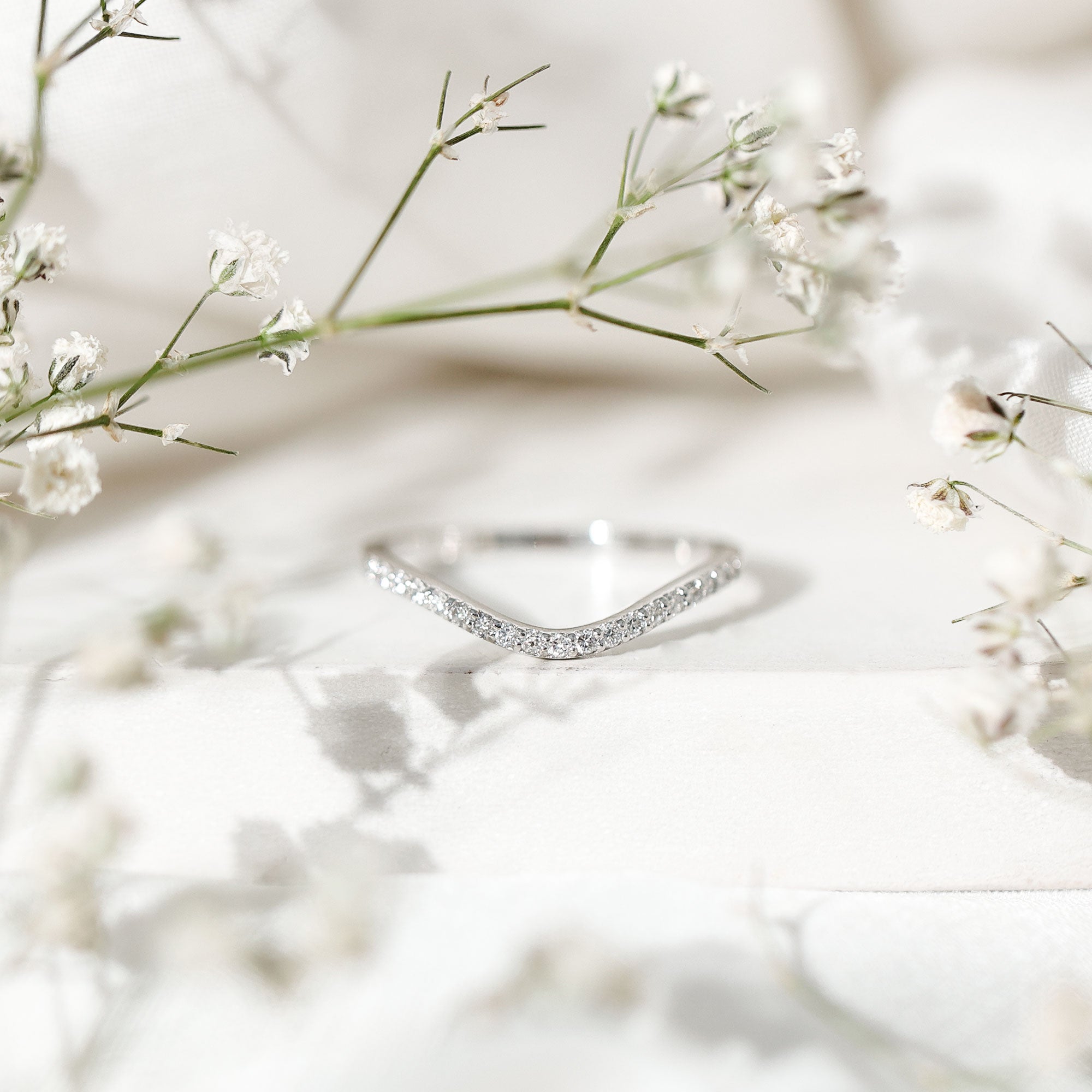 White gold contour diamond wedding band with a soft chevron curve, photographed on a white pedestal with flowers.
